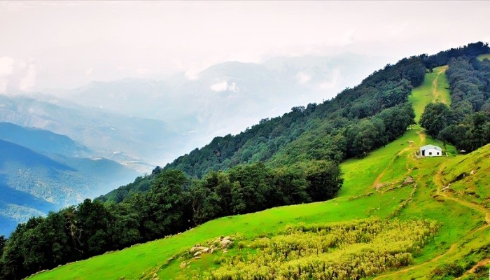 Panoramic view from Nag Tibba viewpoint featuring lush green meadows, dense Himalayan forests, a hillside trail, and misty mountain ranges in Uttarakhand.